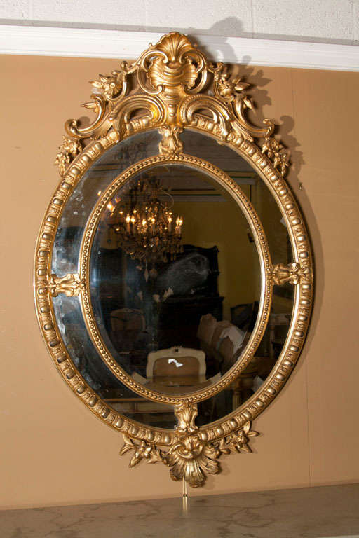 An elegant English Georgian style giltwood mirror, circa 1920s, the oval mirror with double annulated frame, center with a finely-carved crest of scrolls, shells, and flowers.