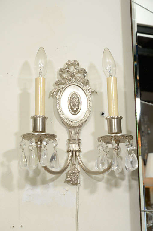 This pair of 1940s Hollywood sconces were realized in the United States, circa 1940. They feature brushed nickel bodies offering cylindrical necks; circular bobeches with foliate detailing; and curved and fluted arms that attach to the reeded