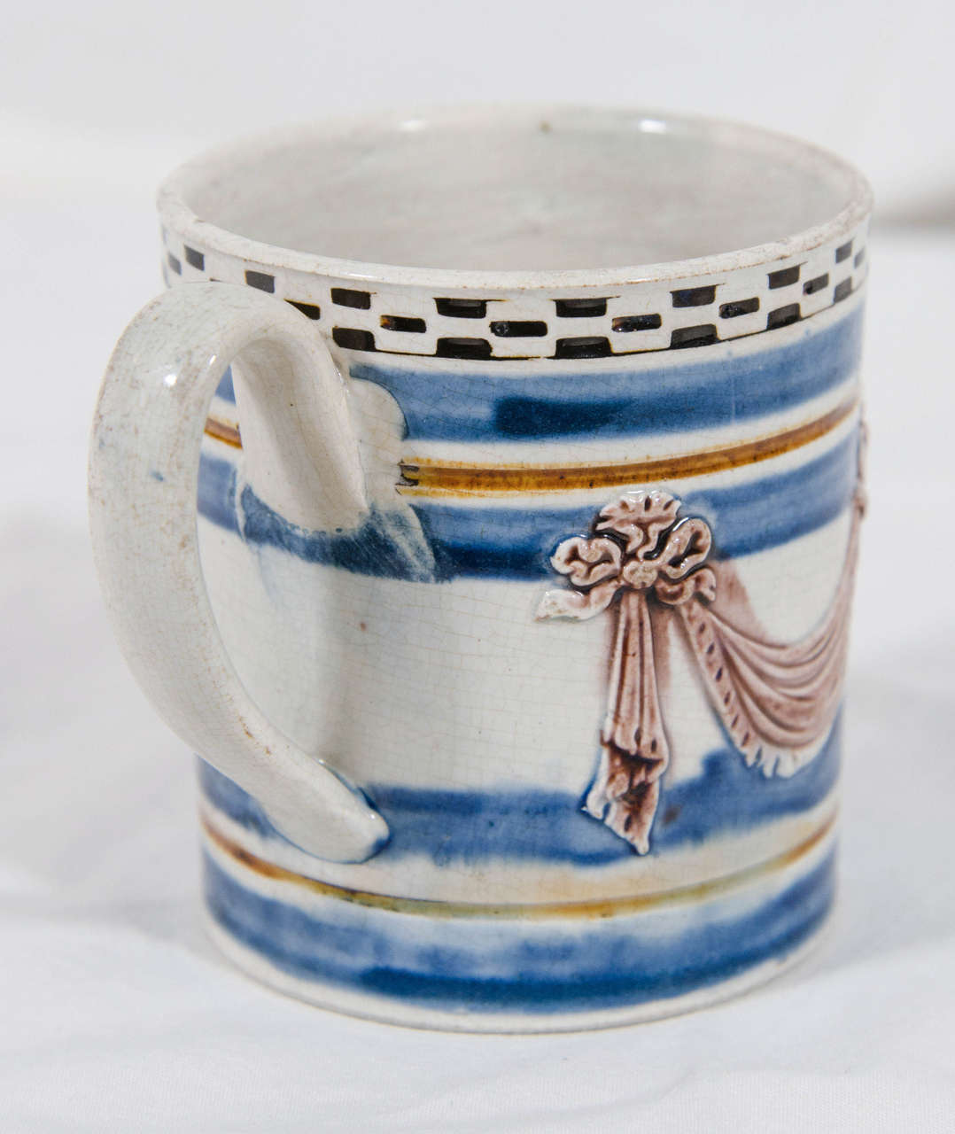 A small creamware mochaware child's mug with a lively design of black and white rouletting, manganese draped swags, and bands of blue and brown.