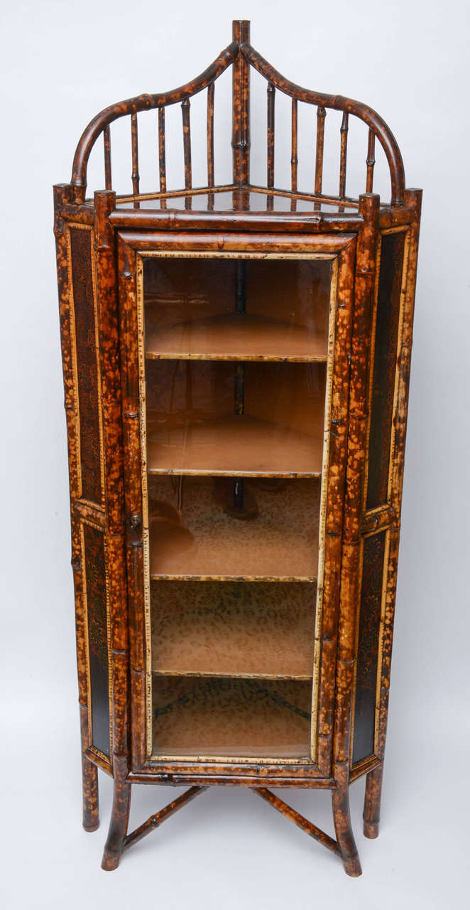 19th century, English. Bamboo corner cabinet with bent and slatted bamboo accents at top surrounding gold, red and green hand painted florals on top on black ground. Matching painted accents on side panels. Hinged glazed door opens to five shelves