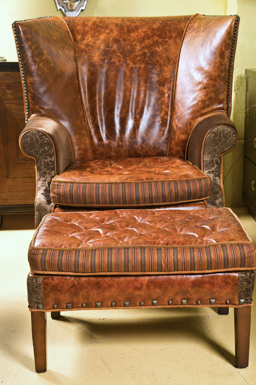 A signed Jeff Zimmerman wingback bergere chair and matching ottoman, circa 1970s-80s, both upholstered in distressed leather decorated with nail heads, cushioned seat, rolled arms, raised on squared tapering legs. 

Ottoman Dimensions: 16.5 Height