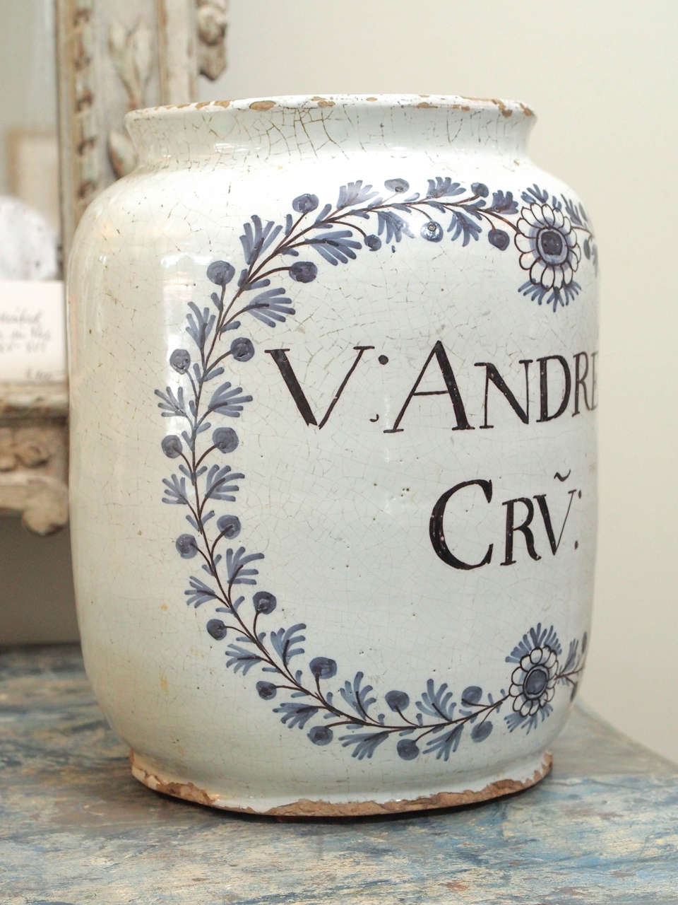 A large tin glazed apothecary style jar in blue and white, paint decorated with a wreath of leaves and berries, and encircling black print label.