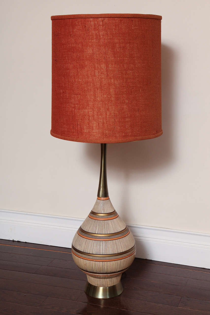 Glazed in cream, brown, orange, black and metallic gold with the neck and base wrapped in metal. Retaining its original deep orange woven shade and signed and dated 1961 at base of lamp. Lamp base itself measures 25 1/2
