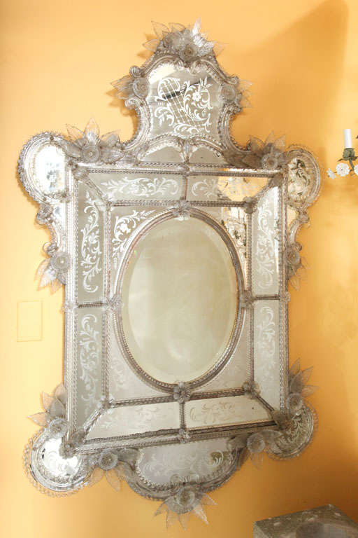 An Italian Venetian glass mirror with beveled oval center looking glass surrounded by sections of etched glass and shaped rods secured by rosettes, the rounded extended corners framed with sections of Murano glass and surmounted by crest with Murano