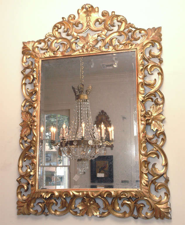 An open work carved gilded frame with a simple crest with bell flowers and stylized leaves and rinceaux overall, surrounding a beveled mirror.