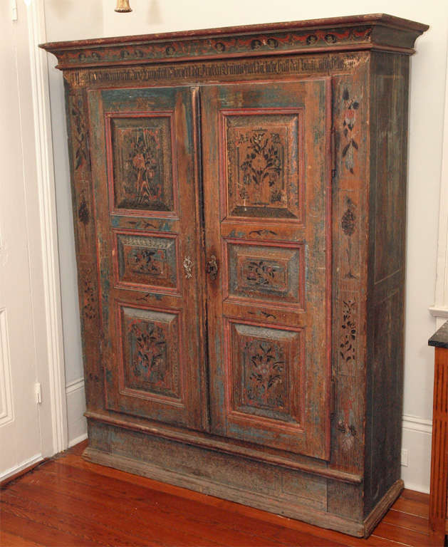 An armoire with paneled doors and well molded cornice.  Retaining the original paint, now worn, but of hues of reds and blues, and depicting flowers in vases, and birds perched atop topiary trees.  Beneath the cornice the paint is of lettering and