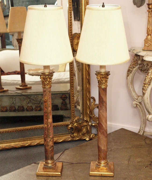 Near pair of faux marble Corinthian columns, not matching but similar, re-purposed as lamps. One of the columns has a filigree design painted in gold on one side. A beeswax sleeve surmounted on the column serves as a base for harp and bulb. The