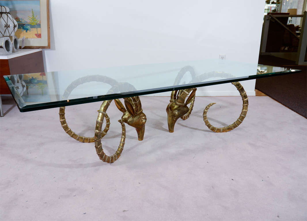 A coffee or cocktail table composed of two bases and a rectangular beveled edge glass top. Each base is brass and in the form of an Ibex head.