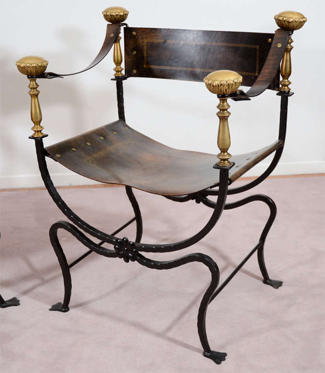 A pair of Curule form chairs composed of an iron frame and leather backs and seats with brass detailing. The leather has a gold box detail with stylized border as well as gold screws joining the leather.<br />
<br />
Reduced from: $4900
