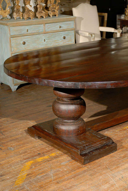 An oval dining room trestle table. This dining room table features an oval top made of several planks raised on a hand-carved trestle base. The two turned legs are joined with a cross stretcher. This oval table is made of reclaimed wood.