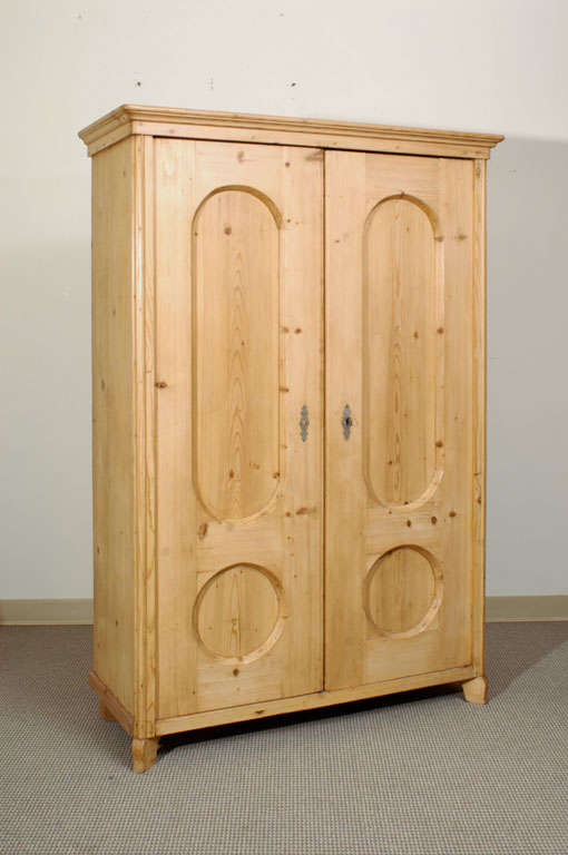 A pine armoire of delightful blonde colour with two doors, each featuring one oval and one round panel. The doors open on the original lock and key to reveal a strip of wooden hanging pegs within.
