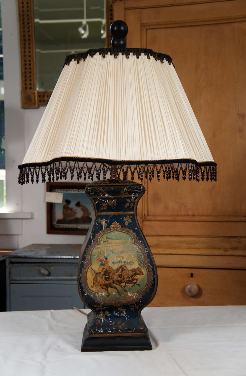 Continental painted tole lamp with allegorical scenes of Roman charioteers on one side and godlike figures fighting it out on
the other on dark blue ground. Some paint loss, but contributes to nice overall look. Topped by off-white ruched lampshade
