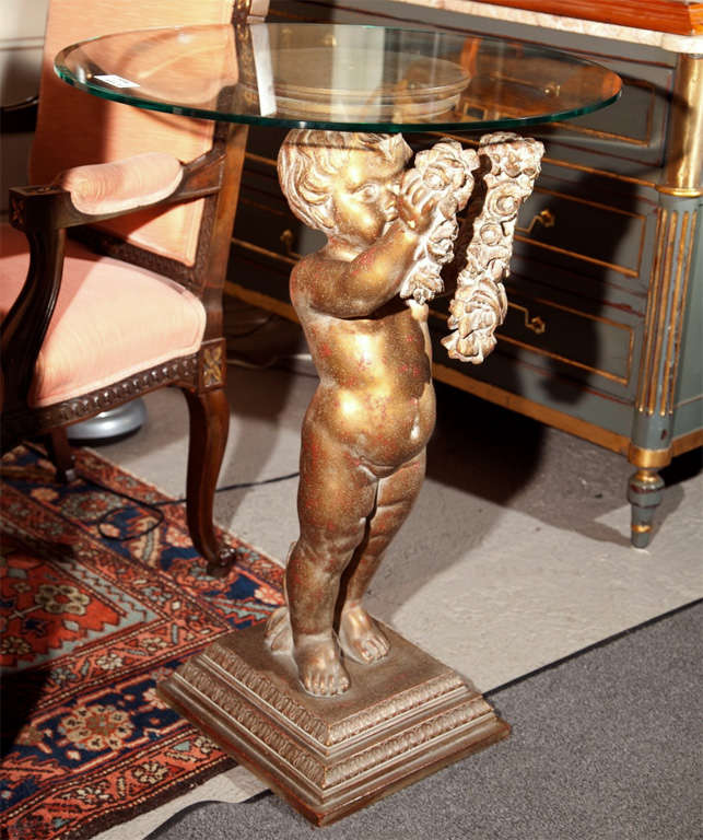 French pedestal table with circular glass top, the elegantly carved base of a cherub holding garlands, raised on squared base.