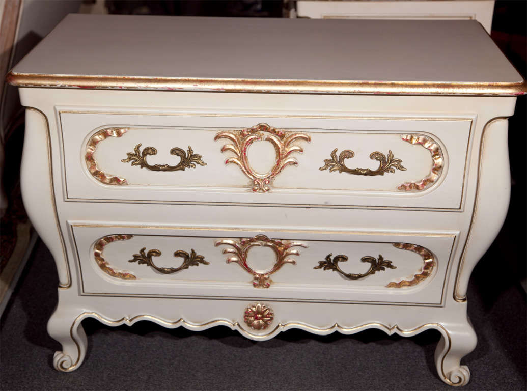 Pair of French, Louis XV style bombe low chests, overall white painted and parcel-gilt, the rectangular top over a conforming case fitted with two drawers, raised on scrolled legs.