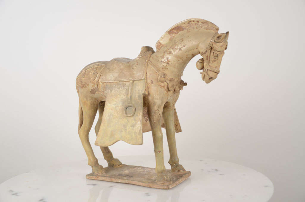 A Chinese glazed potery model of a static horse, with flared saddlecloth, the harness fittings elaborately moulded and with a bell beneath its head, covered in a crackled cream-coloured glaze. SUI Dynasty, A.D. 589-618 caparisoned and with traces of