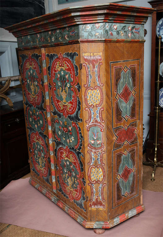 A painted two-door Austrian cabinet, the cornice with faux marbleized paint over floral decorated trees, the two doors with painted rococo cabochons with flowering urns, the canted corners similarly decorated, the side with strapwork painted faux