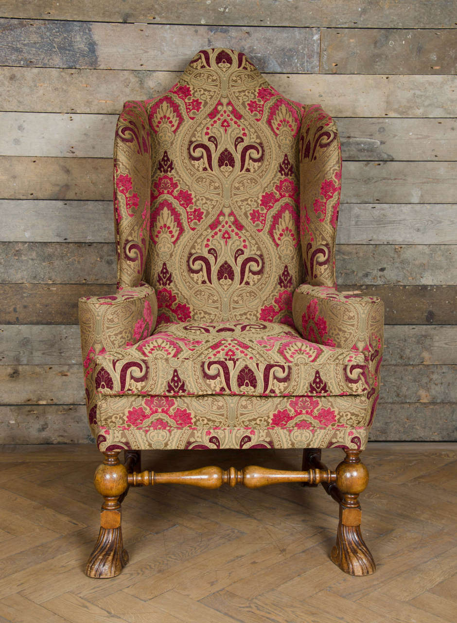A majestic and grand antique armchair in the William and Mary style. This large, high-backed armchair has elegantly scrolling arms and scalloped wings. The base has turned legs and stretchers over finely carved 'Spanish' feet. The chair has been