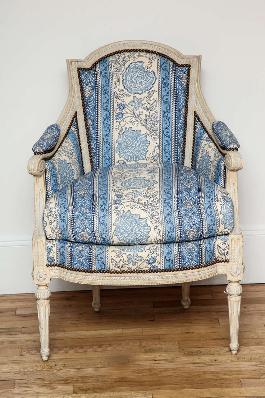 Louis XVI Grey-Painted Bergere 
Chair has an arched incurved Rectangular padded back, bowed cushion seat in molded carved wood frame, and round fluted legs. Upholstered in a large scale blue striped fabric with floral center fields.