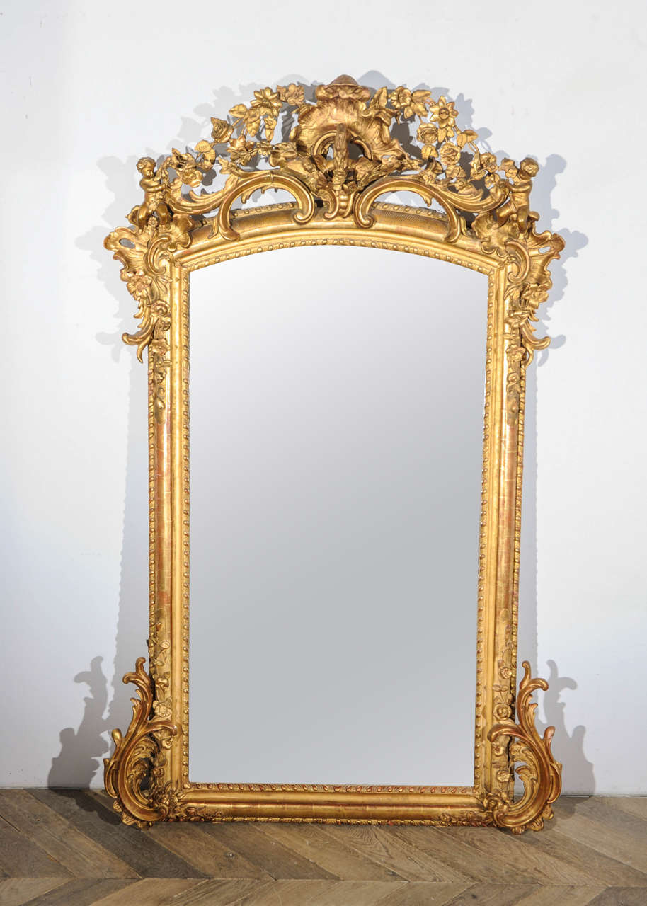A large and richly decorated 19th century French giltwood and gesso wall or mantel mirror in Rococo style, crested with a dragon seated on a coquille, flanked by putti and scrolling acanthus, with floral-festooned shaped frame.