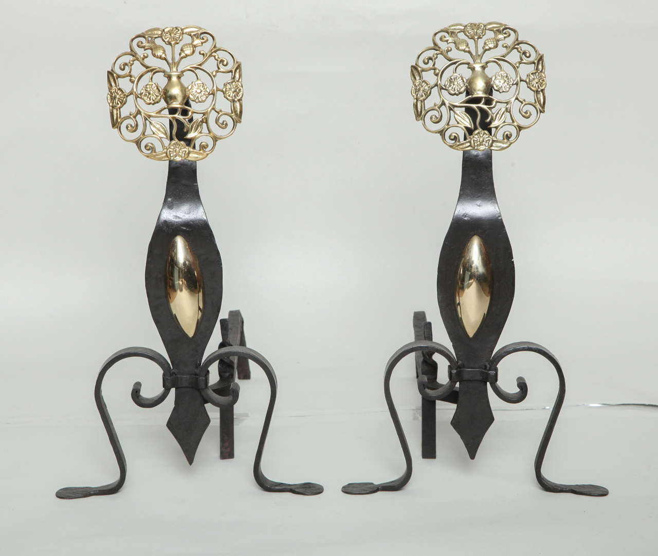 Fine pair of Arts and Crafts period andirons, the filigree brass tops depicting flowering vases, over hand-hammered iron shafts with brass lozenges, standing on scroll feet.
