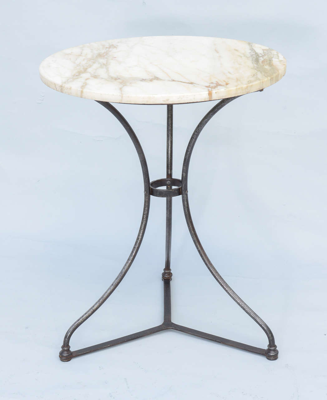 Round bistro table, having white veined marble top, raised on three iron legs connected by a stretcher.