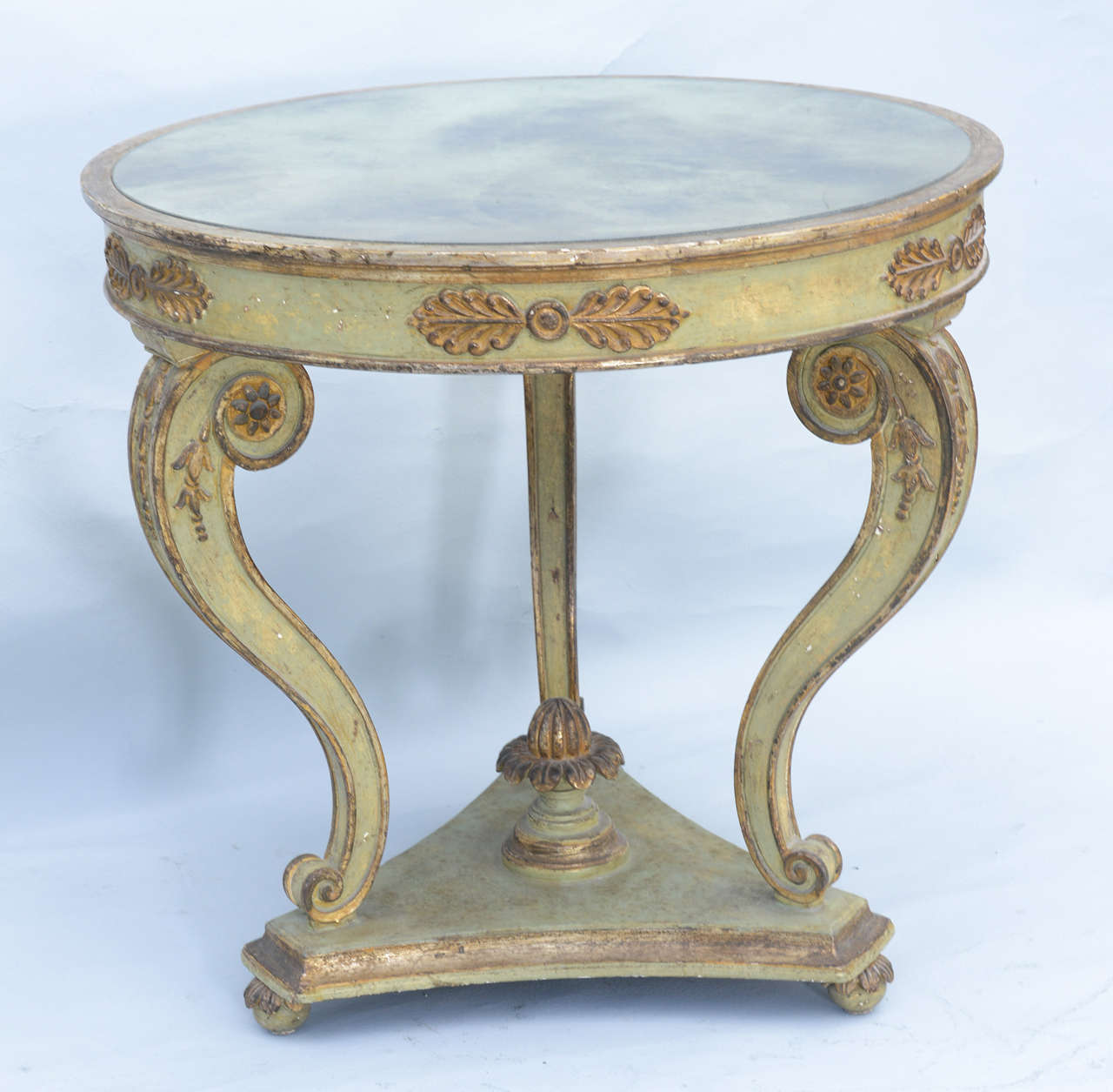 Round, center or end table, painted and parcel gilt, having distressed mirrored top inset in frame on apron outcarved with anthemion, raised on three cabriole legs with rosettes, acanthus knees and scrolling feet, raised on concave tripartite base