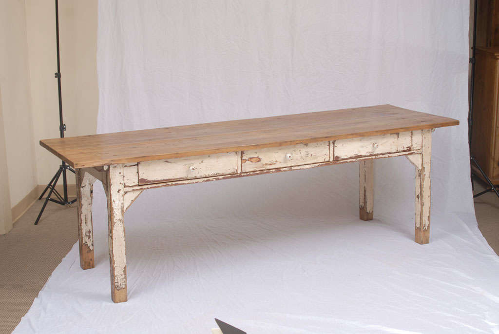 A farmhouse or work table on four straight legs featuring three hand-cut dovetailed drawers with original ceramic knobs on one side. The base is in heavily distressed and crazed white paint.