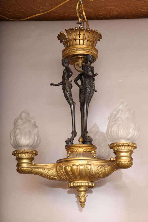 Empire chandelier, of patinated and gilt bronze, originally oil burning; having a central standard made of a trio of monopids, each cast as a well articulated satyr, the mythological creature Pan, on a tripartite bottom holding three flame-shaped
