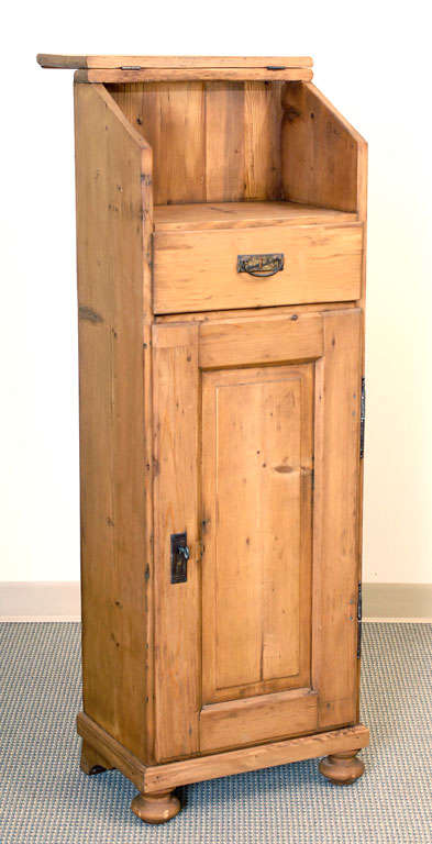 A narrow pine lectern featuring hinged slope-top above notes/bookshelf and single dovetailed drawer. A single raised-panelled door on iron barrel hinges opens 180 degrees to reveal a single shelved locking cupboard.
