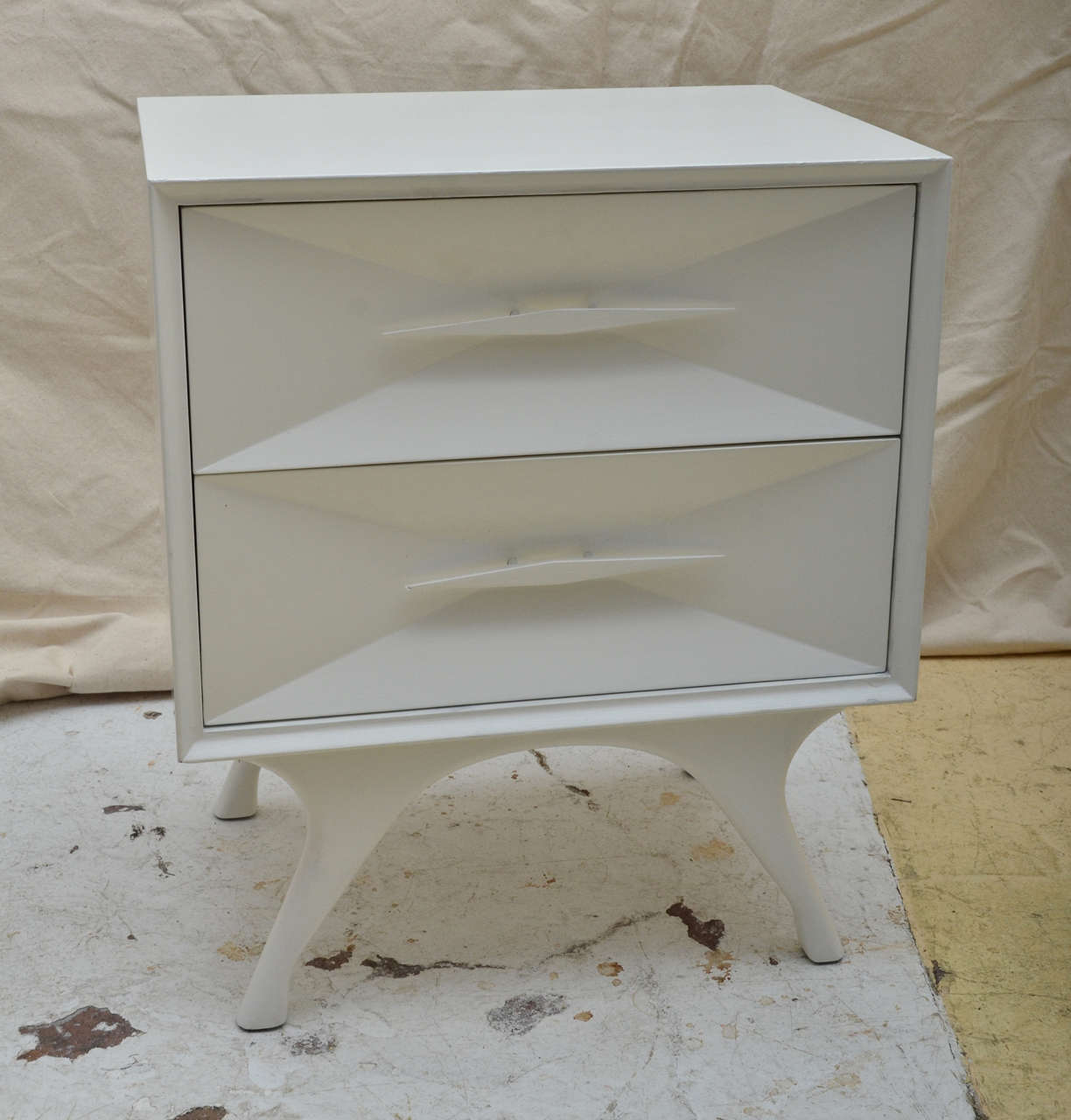 Pair White Painted 1960 American Mid Century Modern Bedside Tables Raised on 4 Splayed Legs. The Drawer Fronts Form An Inverted Pyramid Design, Centered by Narrow wood Pulls. Stamped on Bottom of Drawer Is a Circle With BDB in A Triangular