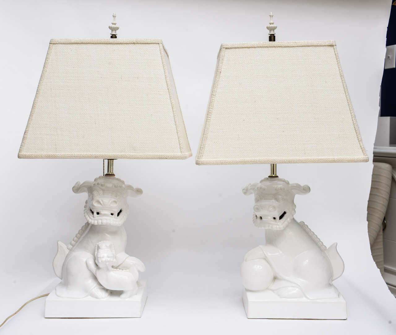 Lovely pair of white ceramic foo dog lamps with custom cream burlap shades. Original bakelite chinoiserie finials included.
Minor knick on one lamp which restorer identifies as too small to repair--does not distract from the beauty of these light