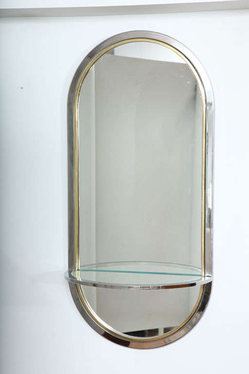 An elegant elongated oval chrome and brass framed mirror with glass shelf. In the style of Paul Evans.