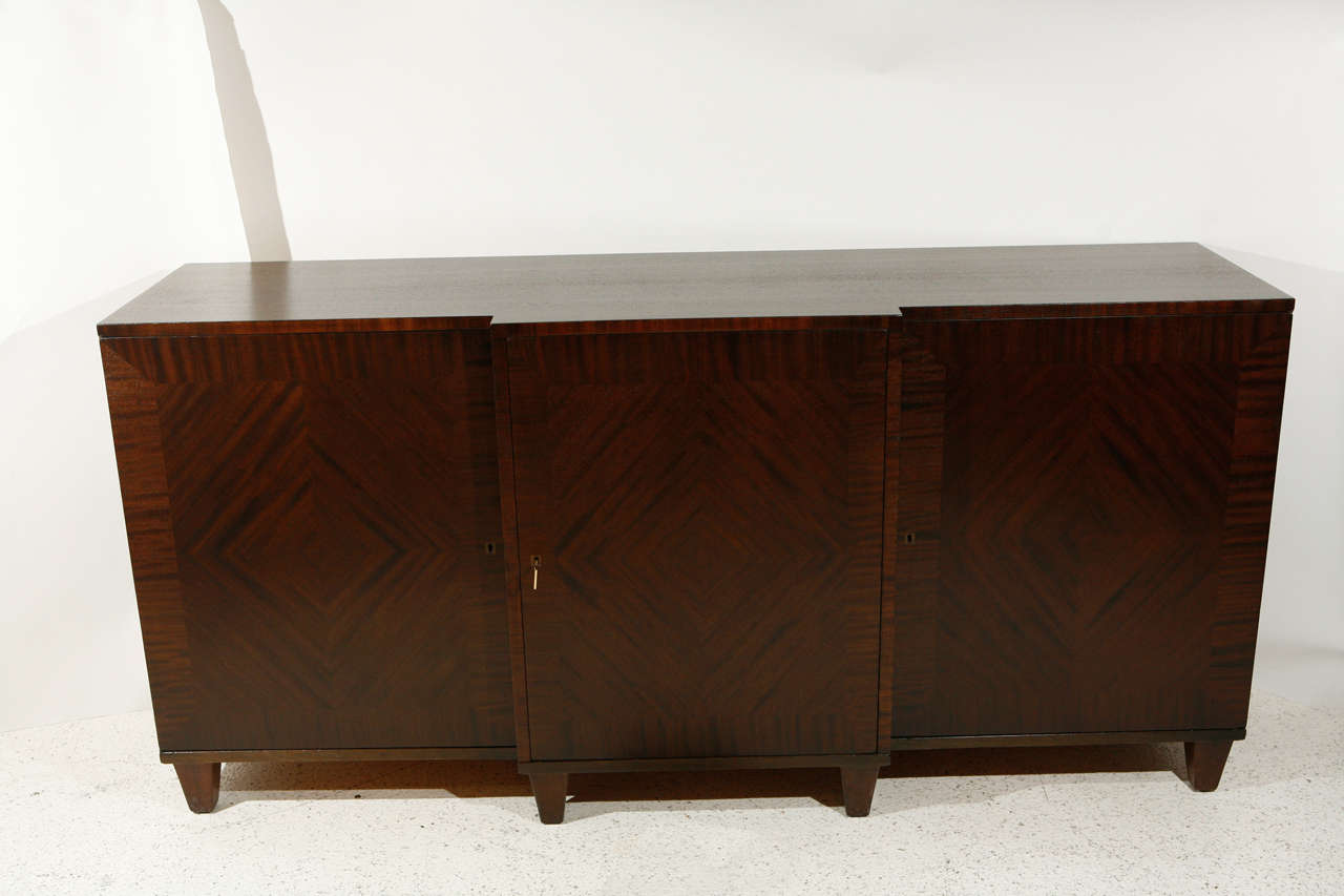 A handsome three-door buffet veneered in ribbon mahogany with set-back back side sections. The cabinet features diamond matched veneers on the front of all three doors and a nice channel reveal at the base of the cabinet. The stepped out center
