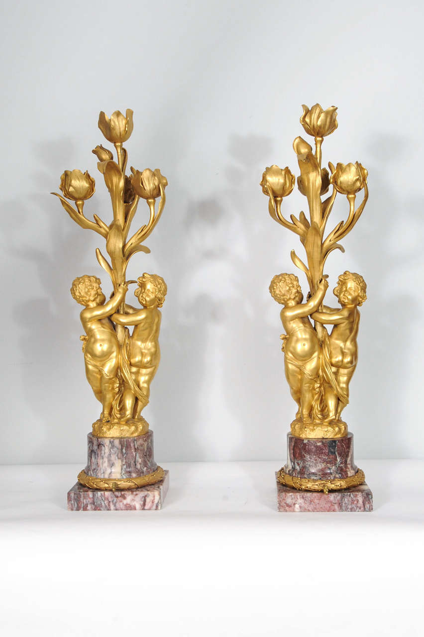 Both these lamps have two finely cast and chiseled gilt bronze putti holding a floral stem with leaves, the three lower buds with lamp-fittings, the highest lacking the stand for a shade and fourth lamp, marble base with original turn-switch and at