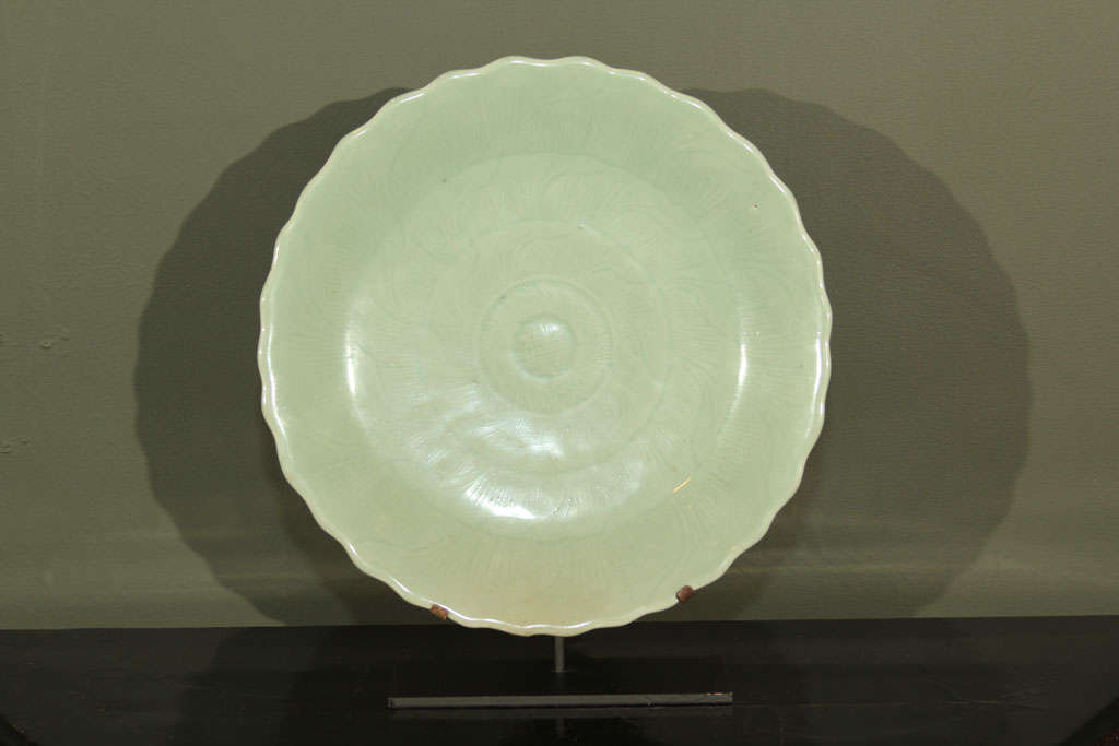 Japanese porcelain charger bowl with green-blue celadon glaze. Of circular form with scalloped rim and slightly raised foot. The surface decorated with incised stylized flower head with concentric rings of petals. The glazed foot with apocryphal