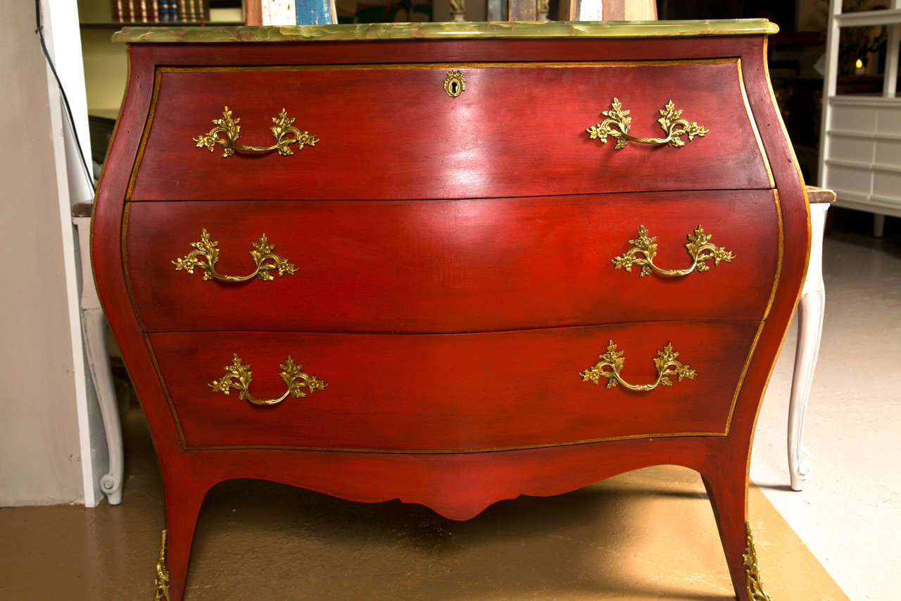 French Louis XV style bombe chest of drawers, the green onyx marble atop a red distress painted bombe case opening to a drop-front writing surface and cubby holes, raised on splayed legs decorated with bronze mounted sabots.