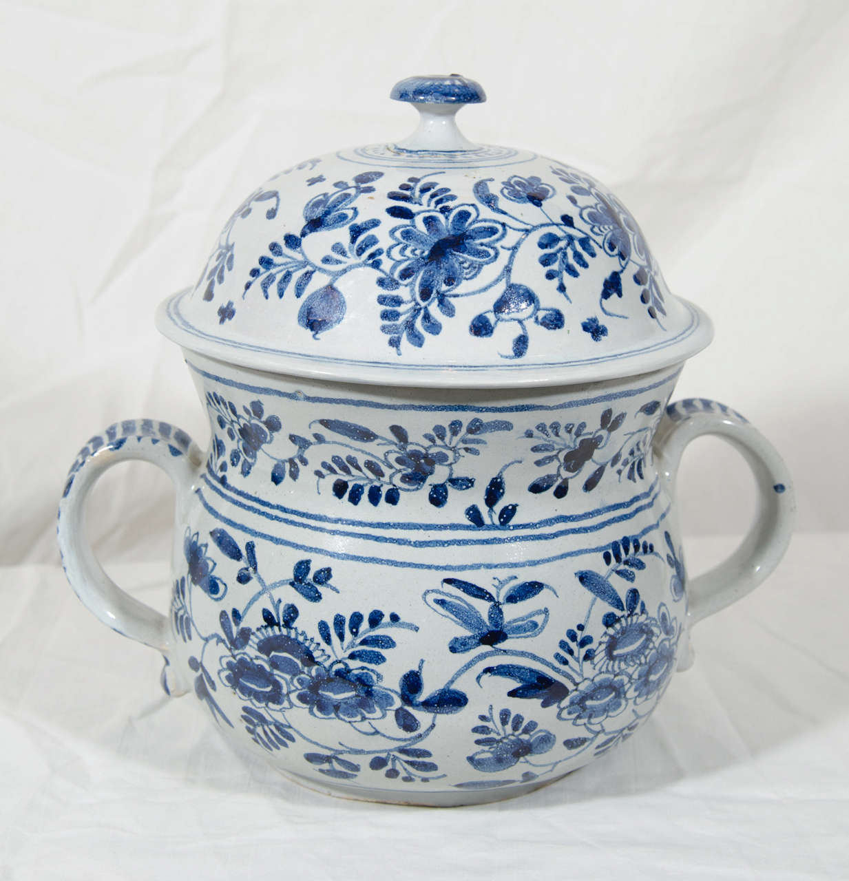 A blue and white Delft posset pot and cover decorated in underglaze blue with bands of scrolling flowers. The cylindrical body with a flat shoulder and a short wide neck, the curved spout and strap handles with dashes in blue, and a domed cover with