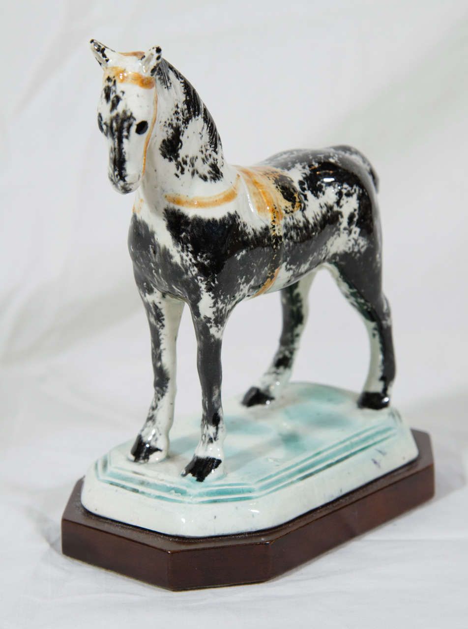 An 18th century Pearlware pottery racehorse dappled gray, with an orange saddle blanket, and orange straps. He stands on a green glazed base with one back foot forward as if he was caught in motion (the Pearlware base supported by a later wood