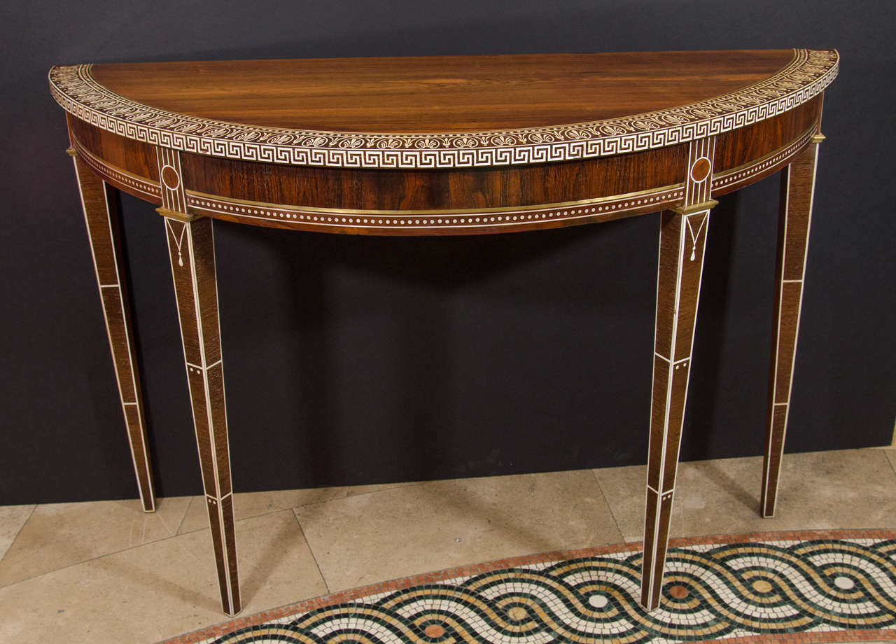 A fine pair of Art Deco rosewood demilune consoles with anthemion and Greek key banding, brass inlay and trim on square tapered and inlaid legs.