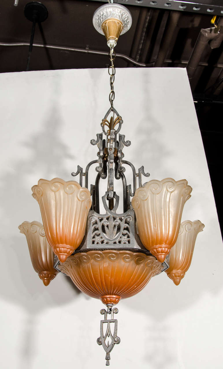 Beautiful Art Deco chandelier comprised of wrought iron and spelter metal frame with incised skyscraper designs and hand applied gilt accents. The chandelier has molded frosted relief glass shades and center dome in gradient hues of copper and