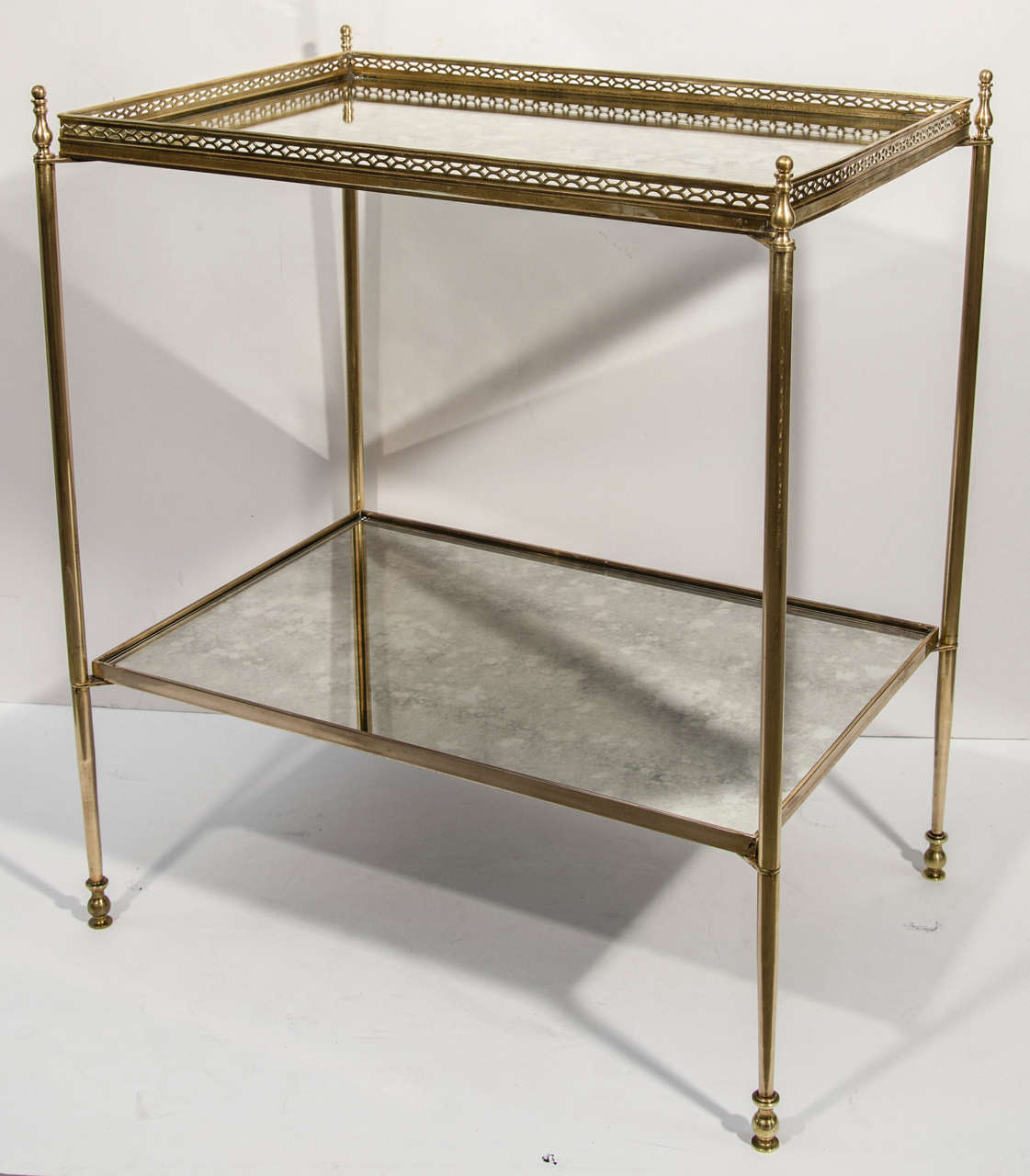 Classical two tier side table in brass with smokey antique mirrored tops. The table features a brass gallery along the top tier with pierced filigree designs, and has elegant cylinder legs with stylized urn form feet.