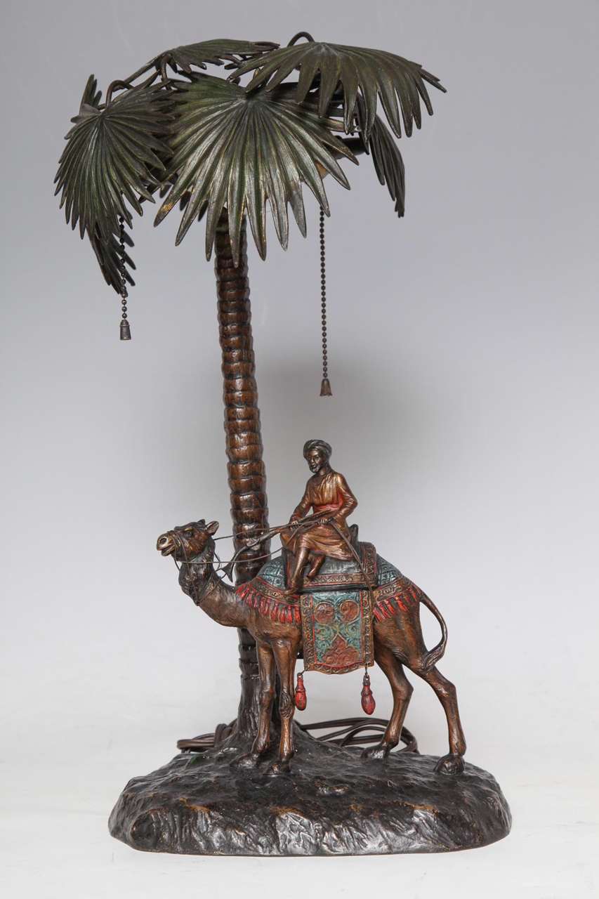 A fine Viennese cold painted bronze orientalist lamp attributed to Bergman of an Arab man riding a camel. The elegantly dressed man sits side saddle on the adorned camel's hump. Both are brightly dressed beneath the towering palm fronds, which