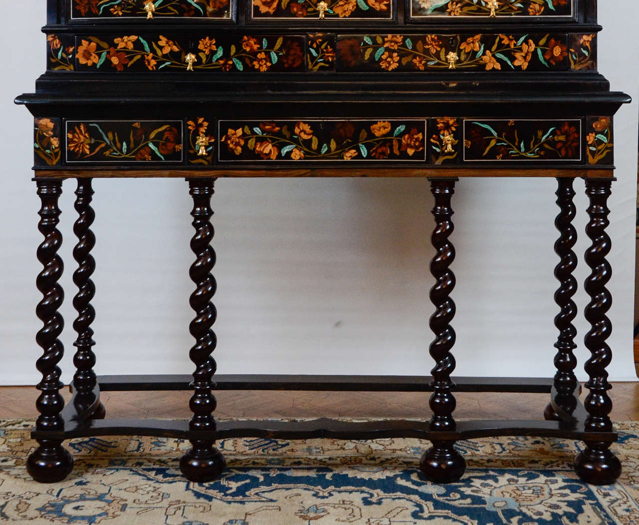The cabinet fitted with 14 drawers and a door on a stand with 
six twist-turned columns joined by a stretcher and above a big drawer. The marquetry of birds, flowers and vases is very luxurious with many different woods, ebony and bone stained