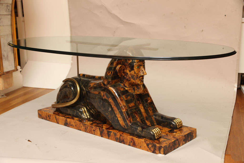A contemporary coffee table with base in the form of a sphinx, the recumbent figure with faux tortoise shell and amber colored veneer. The figure resting on a rectangular plinth, and the head supporting an X shaped bronze frame beneath oval glass
