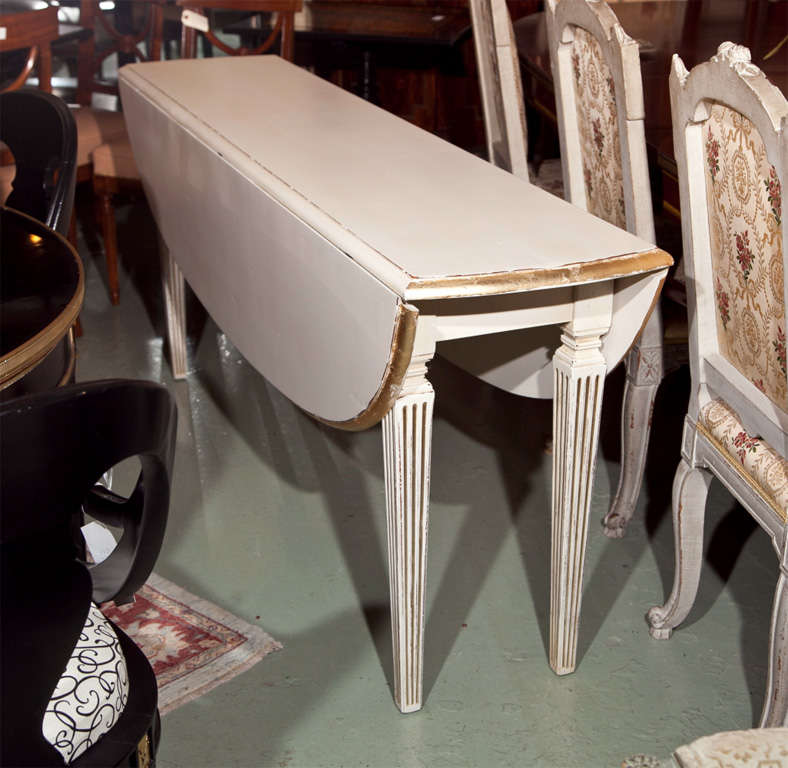 White painted and parcel-gilt drop-leaf dining table, 20th century, the console table with two demilune leaves , raised on squared fluted legs, opens up to an oval small dining table. <br />
Measures 16