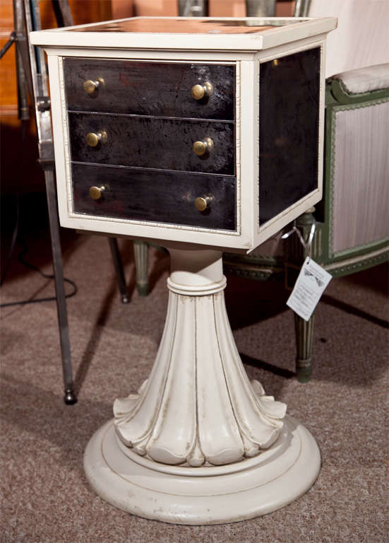 An intriguing Hollywood Regency style jewelry stand, overall white distress painted, the flip-top milar-veneered top of a small conforming case of drawers supported by a plume pedestal, raised on a circular base.