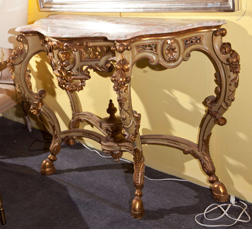 French Rococo style console table, circa 1940, the pink/white serpentine shaped marble atop an ornately carved painted and parcel-gilt base, decorated with foliate scrolls, joint by a stretcher centered by an urn finial.