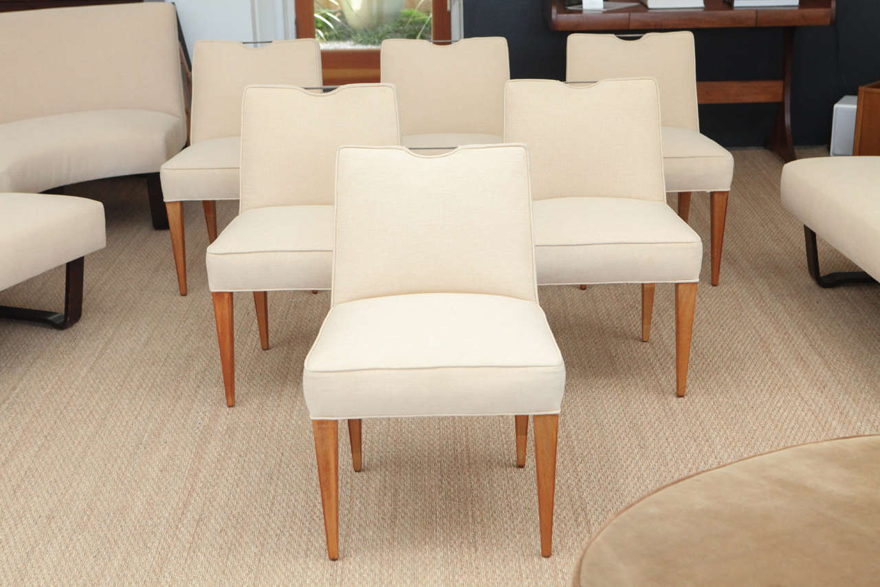 Newly upholstered in a slightly textured ivory fabric, these six armless Dunbar dining chairs are stunning simplicity. The tapered legs are a honey-colored hardwood and the small horizontal steel bar accent in the center of each chair adds the