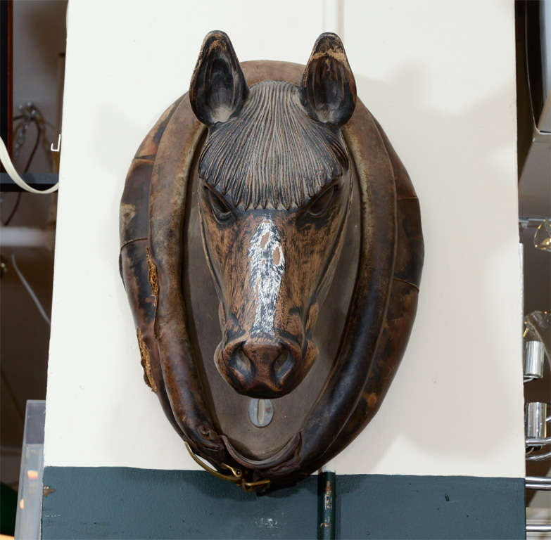 A wall-mounted carved wooden horse head trade sign mounted to a leather base with brass saddle hardware detailing on the bottom.