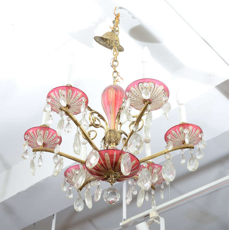 A chandelier with clear and cranberry glass detailing adorned with hanging crystals. The brass frame has scrolling acanthus leaf details.